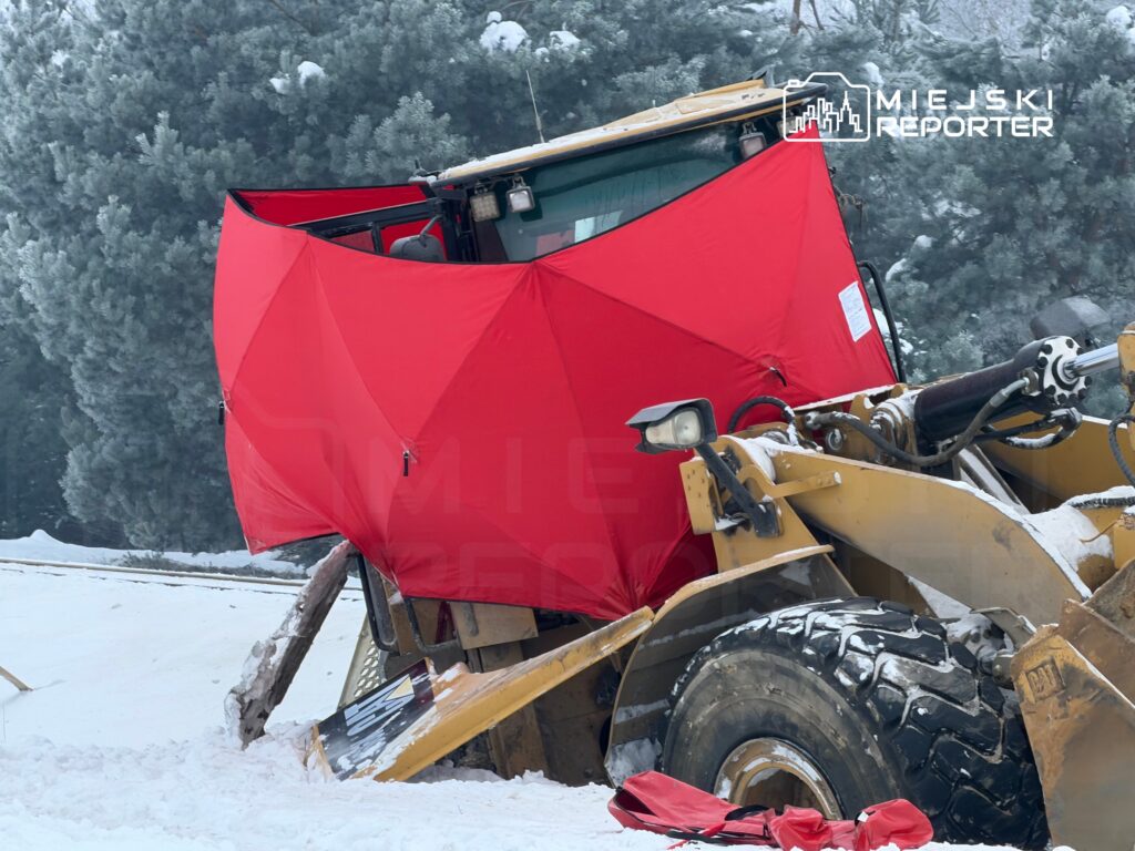 Ładowarka z czerwoną osłoną stoi na zaśnieżonym terenie w otoczeniu drzew.