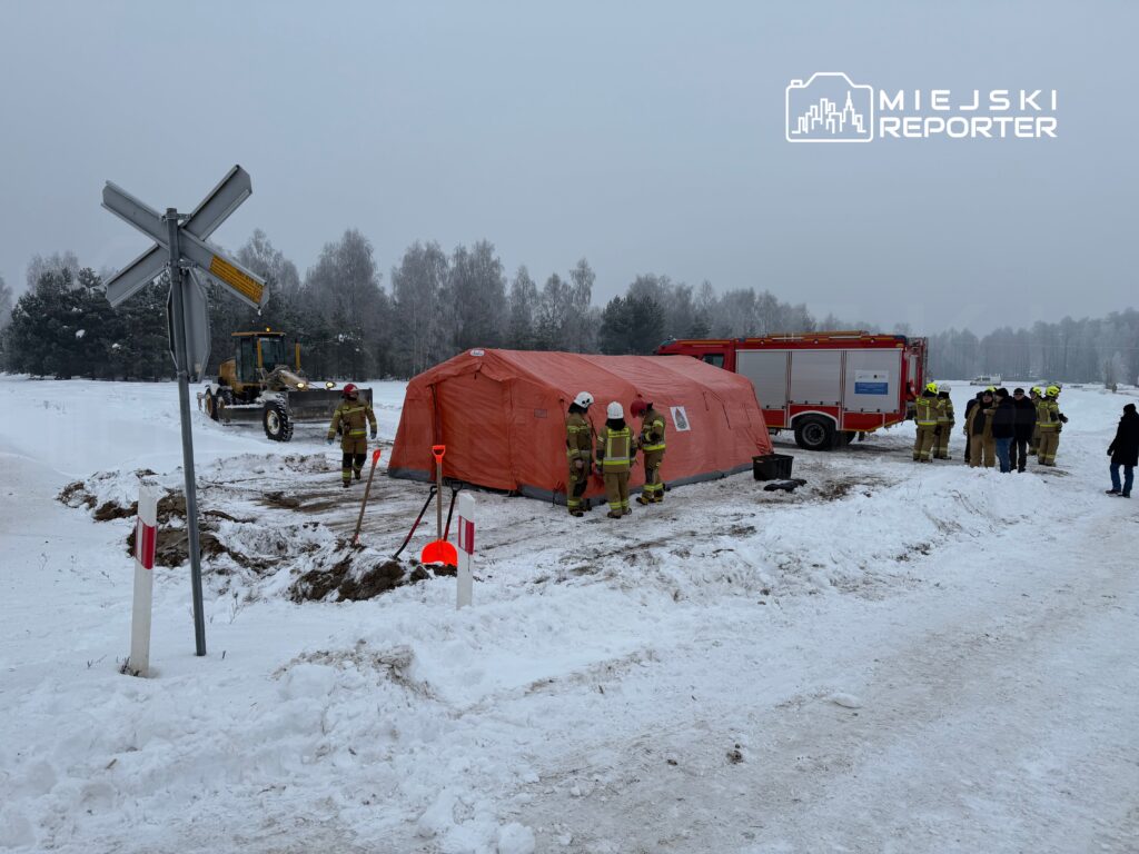 Grupa strażaków pracuje przy pomarańczowym namiocie w zasypanym śniegiem terenie, obok wozu strażackiego.