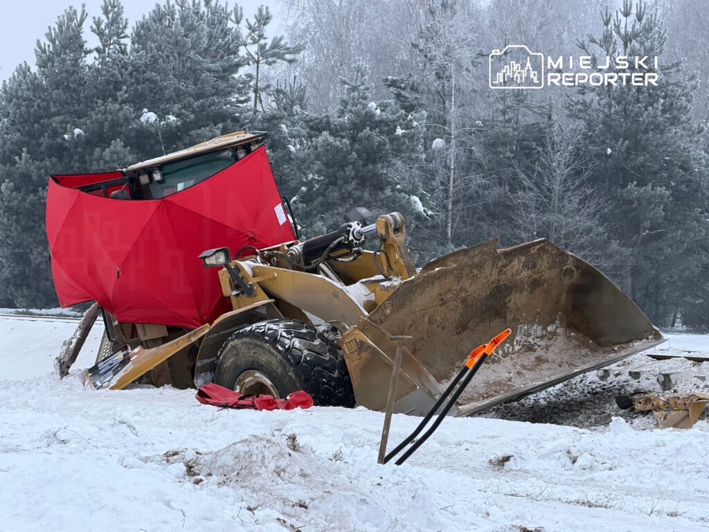 Maszyna budowlana z czerwoną osłoną stoi na zaśnieżonym terenie, otoczona drzewami.