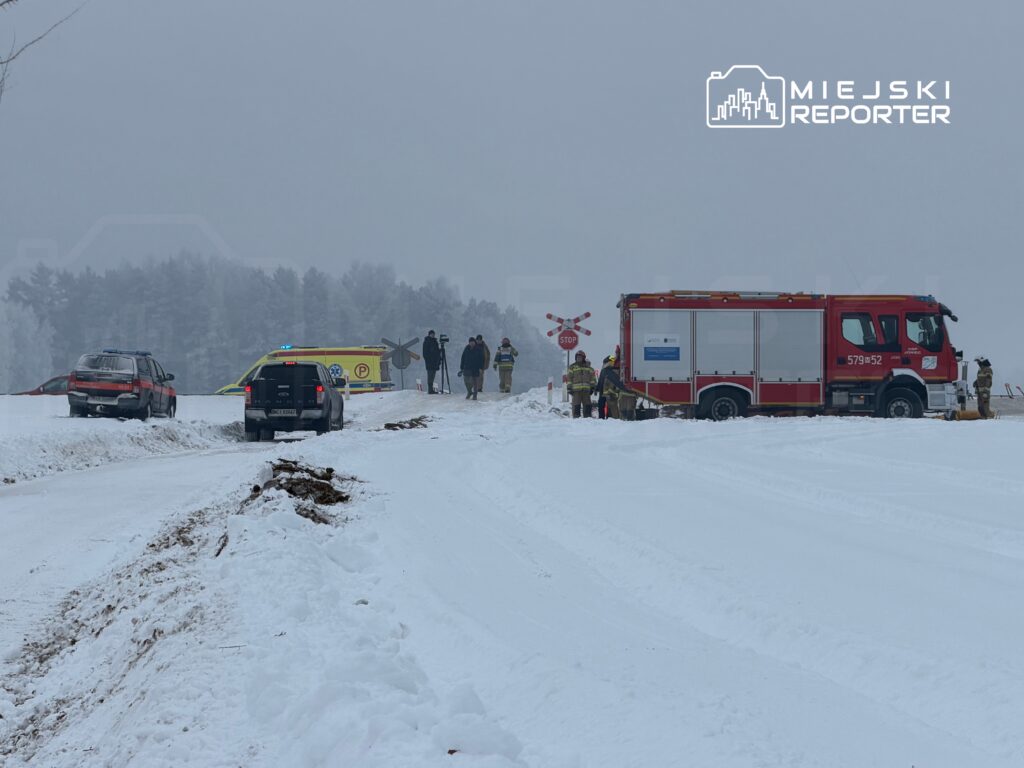 Na zaśnieżonej drodze stoją pojazdy służb ratunkowych, a ratownicy rozmawiają przy wozie strażackim.