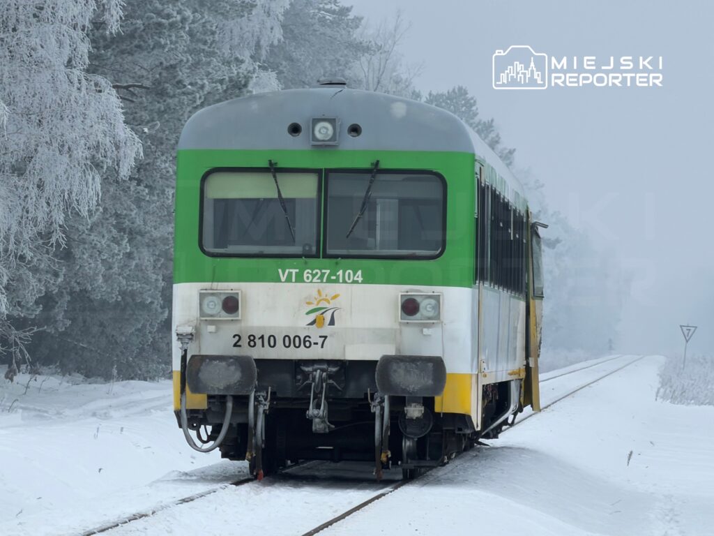 Pociąg osobowy z zieloną i białą karoserią stoi na zaśnieżonym torowisku w otoczeniu zamarzniętych drzew.