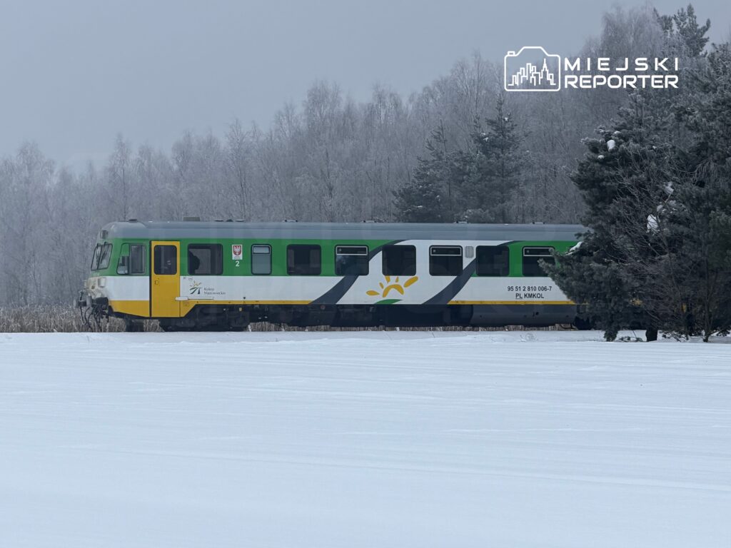 Zielono-żółty pociąg osobowy stojący na zaśnieżonym terenie, w otoczeniu drzew pokrytych szronem.