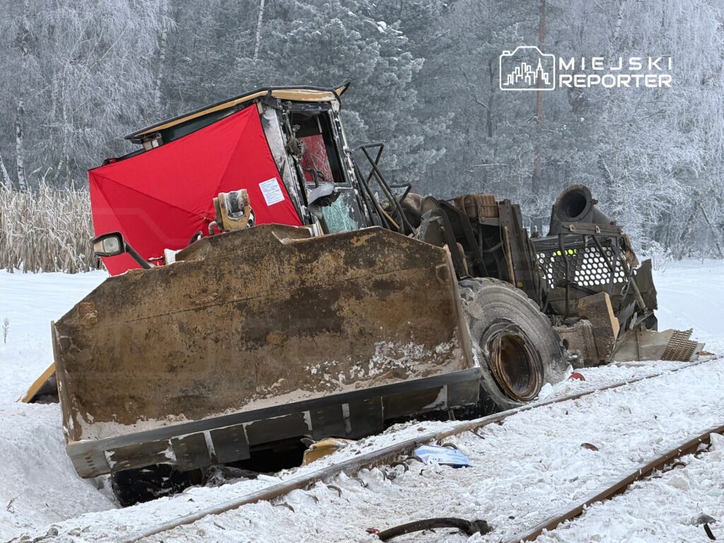 Zniszczony ciągnik gąsienicowy leży na boku w zaśnieżonym terenie, obok torów kolejowych, w otoczeniu drzew.