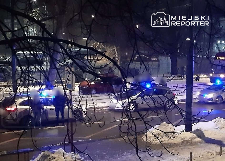 Grupa policjantów rozmawia obok radiowozu na ośnieżonej ulicy w nocy, w tle widać dwa uszkodzone samochody.