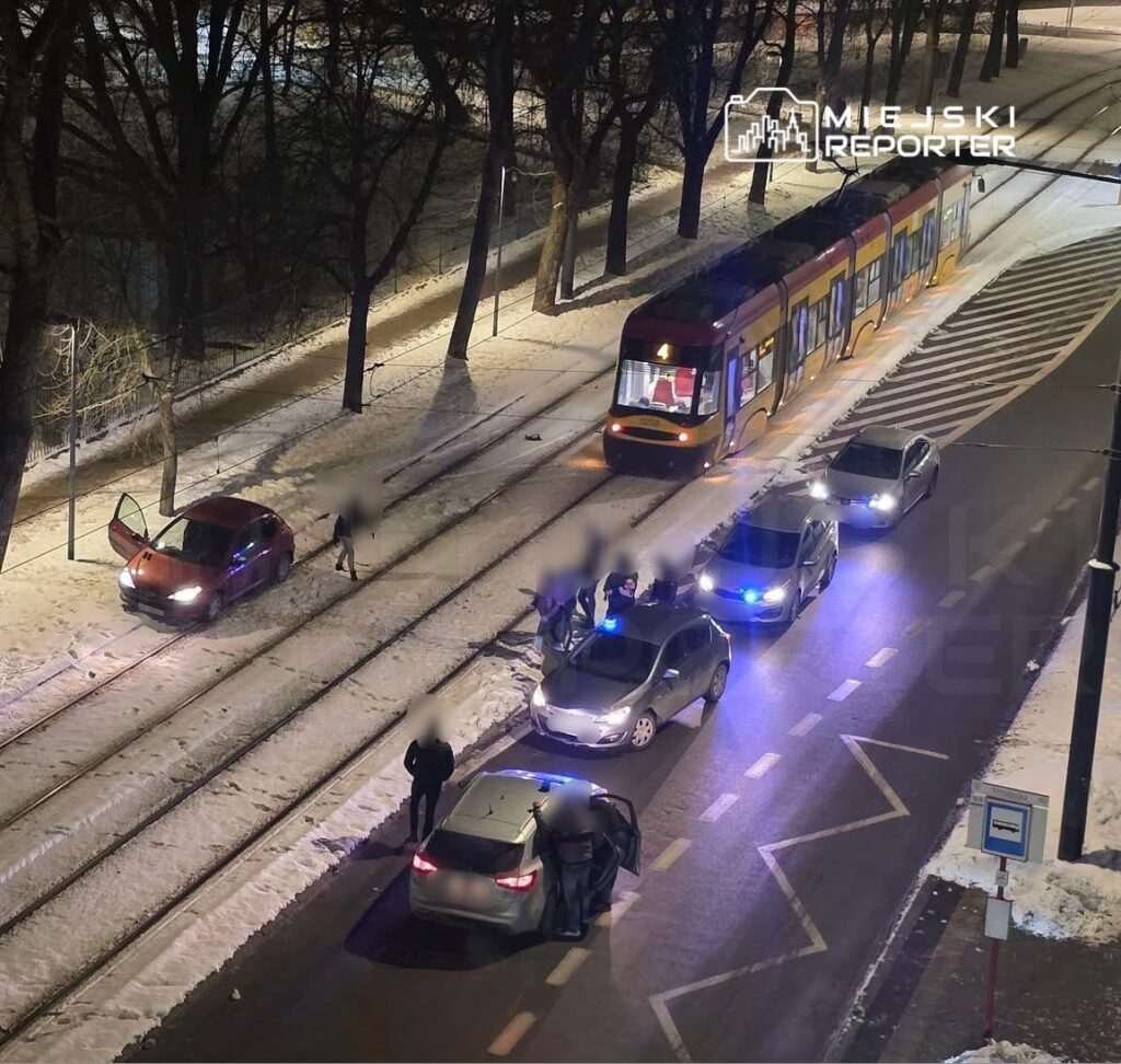 Na ośnieżonej ulicy, przy torach tramwajowych, kilka osób gromadzi się wokół dwóch samochodów, w tym jednego z otwartymi drzwiami. Obok zatrzymany tramwaj.