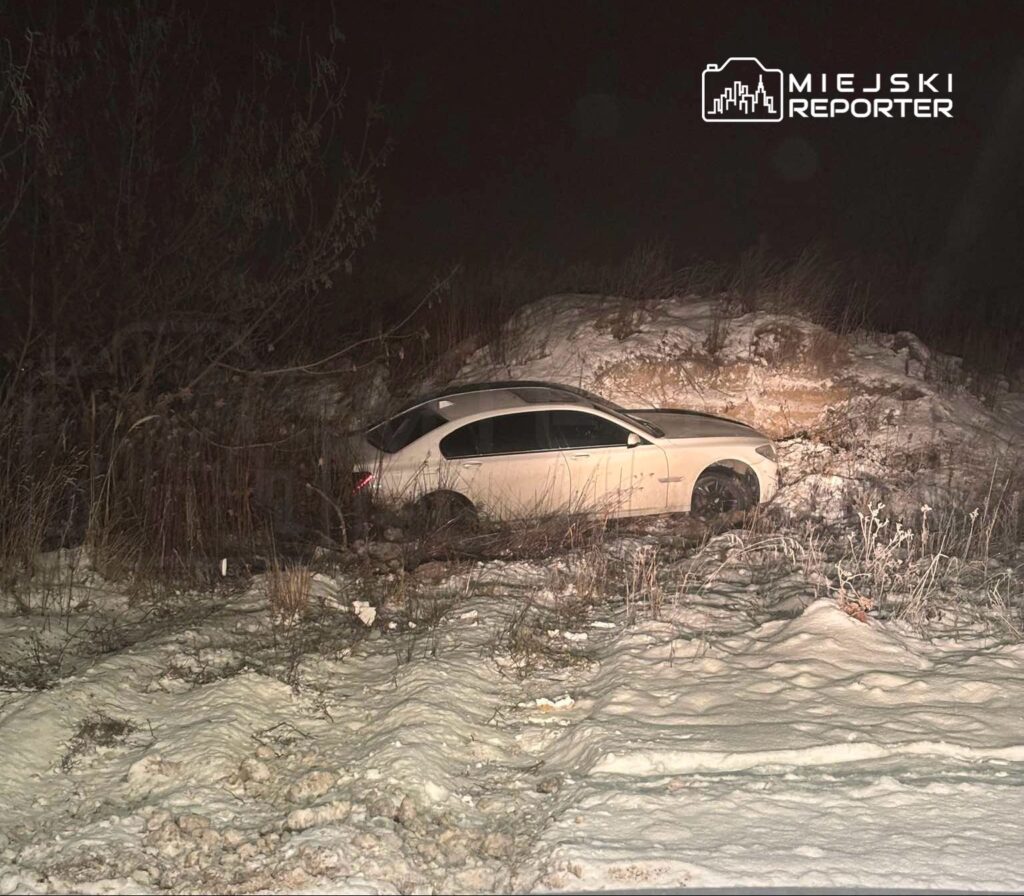 Biały samochód osobowy utknął w śniegu obok zarośli, w ciemnym otoczeniu nocnym.