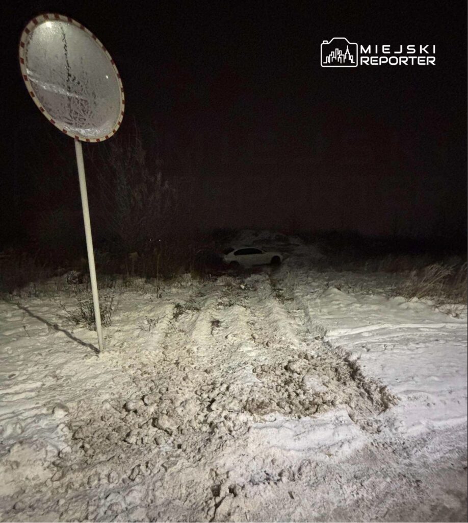 Zasypana śniegiem droga z wyraźnymi śladami opon prowadzi do zaparkowanego w oddali samochodu. W tle stoi znak drogowy.