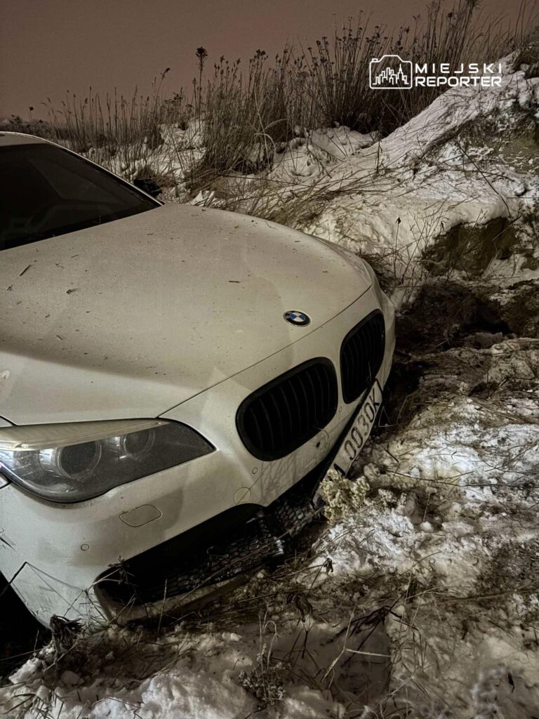 Białe BMW zaparkowane na zaśnieżonym terenie, otoczone wysoką trawą i śniegiem.