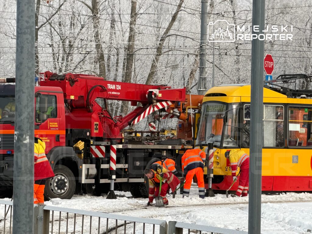 Ekipa ratunkowa w pomarańczowych strojach usuwa śnieg wokół unieruchomionego tramwaju w zimowym otoczeniu.