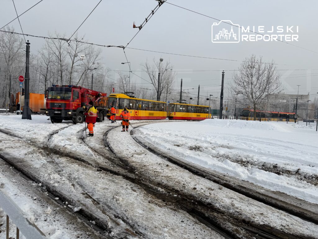 Pracownicy w pomarańczowych uniformach odśnieżają torowisko tramwajowe w zaśnieżonym miejskim otoczeniu.