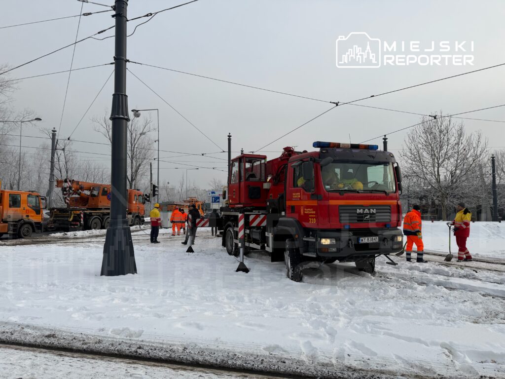 Zespół pracowników w pomarańczowych uniformach odśnieża torowisko tramwajowe w zaśnieżonym otoczeniu.