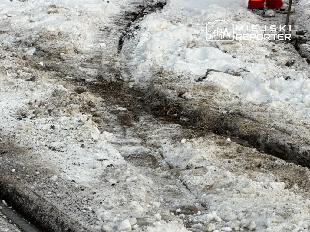 Zabrudzona droga pokryta śniegiem i błotem, z widocznymi śladami opon oraz zaspami po obu stronach.
