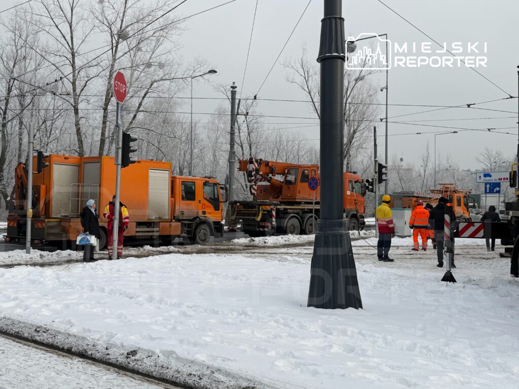 Pracownicy w odblaskowych strojach rozmawiają obok pomarańczowych pojazdów w zasypanym śniegiem otoczeniu.