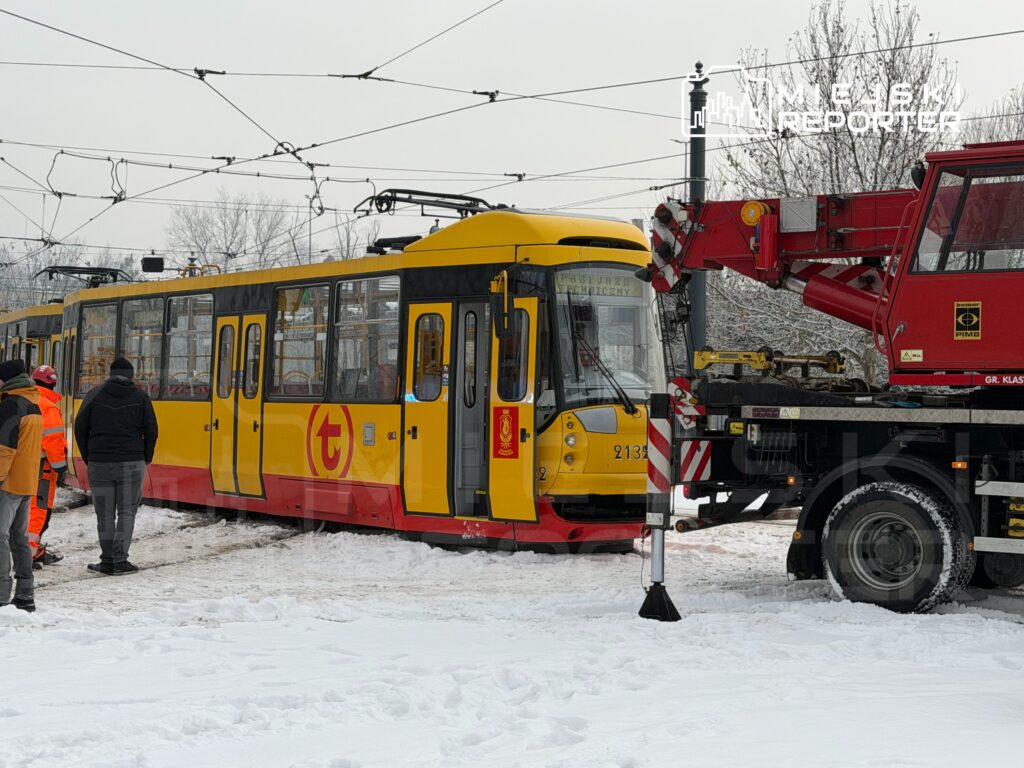 Żółty tramwaj stoi na zaśnieżonym torowisku, obok grupy pracowników w odblaskowych kamizelkach, podczas akcji naprawczej.