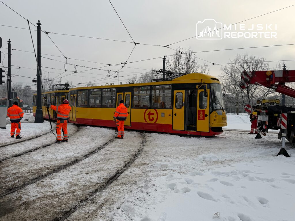 Czterech pracowników w pomarańczowych kurtkach obsługuje żółty tramwaj w śnieżnym otoczeniu.