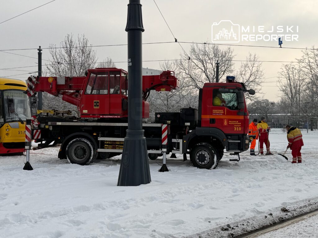 Czerwony pojazd z dźwigiem przy torach tramwajowych, a pracownicy w żółtych kurtkach odśnieżają teren w zimowej scenerii.