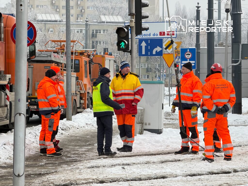 Grupa pracowników w pomarańczowych kurtkach omawia sytuację na zaśnieżonej ulicy, obok wozów strażackich.