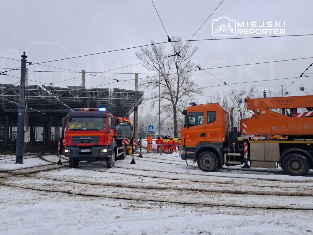 Czerwony wóz strażacki z włączonymi sygnałami świetlnymi oraz pomarańczowy pojazd z podnośnikiem w zaśnieżonym otoczeniu.