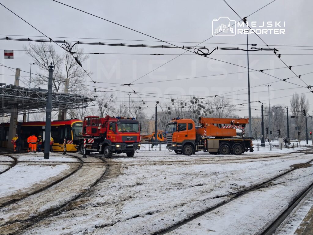 Grupa pracowników w pomarańczowych kamizelkach naprawia tramwaj pod nadzorem wozu strażackiego i pojazdu z podnośnikiem.