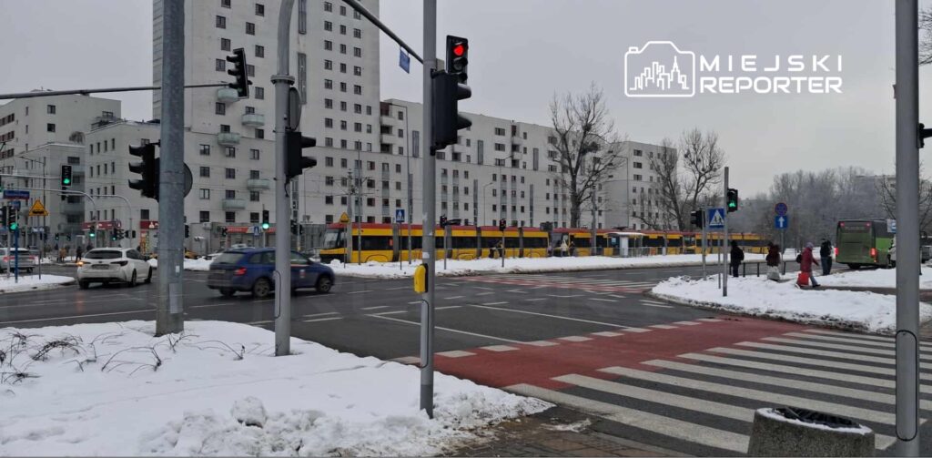 Ruchliwa ulica z tramwajem w tle, samochody na drodze, zima, pokryta śniegiem chodnik oraz sygnalizacja świetlna.