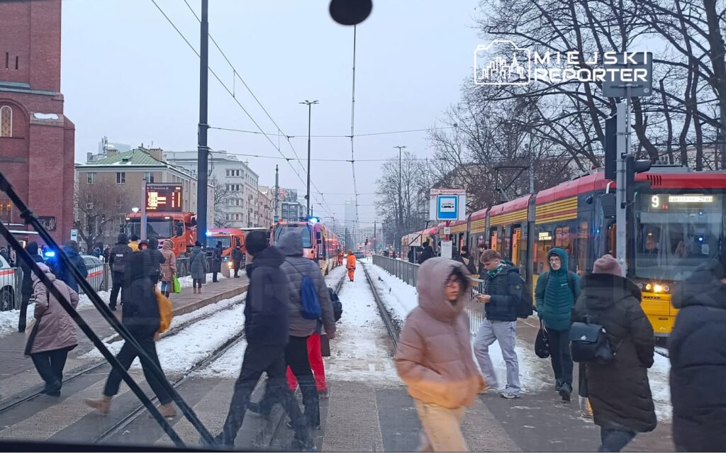 Tłum ludzi czeka na tramwaj na przystanku w zimowej scenerii, obok przejeżdżających pojazdów służb ratunkowych.
