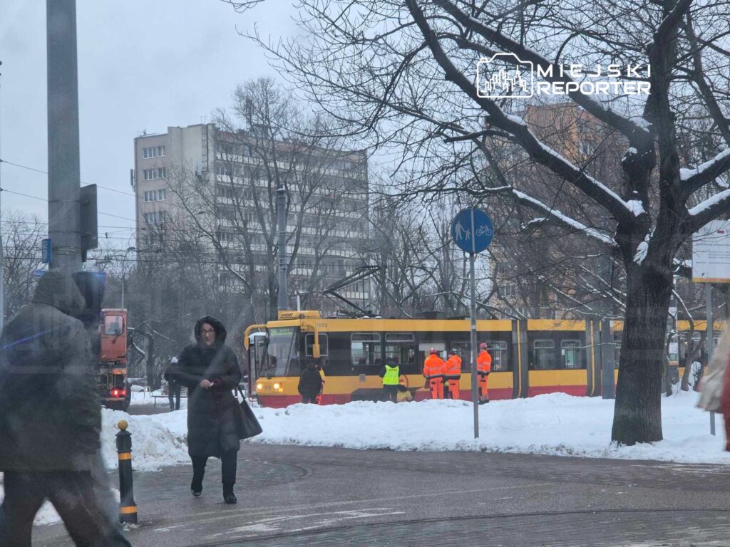 Kobieta w czarnym płaszczu przechodzi obok tramwaju, a pracownicy w pomarańczowych kurtkach pracują na zaśnieżonym przystanku.