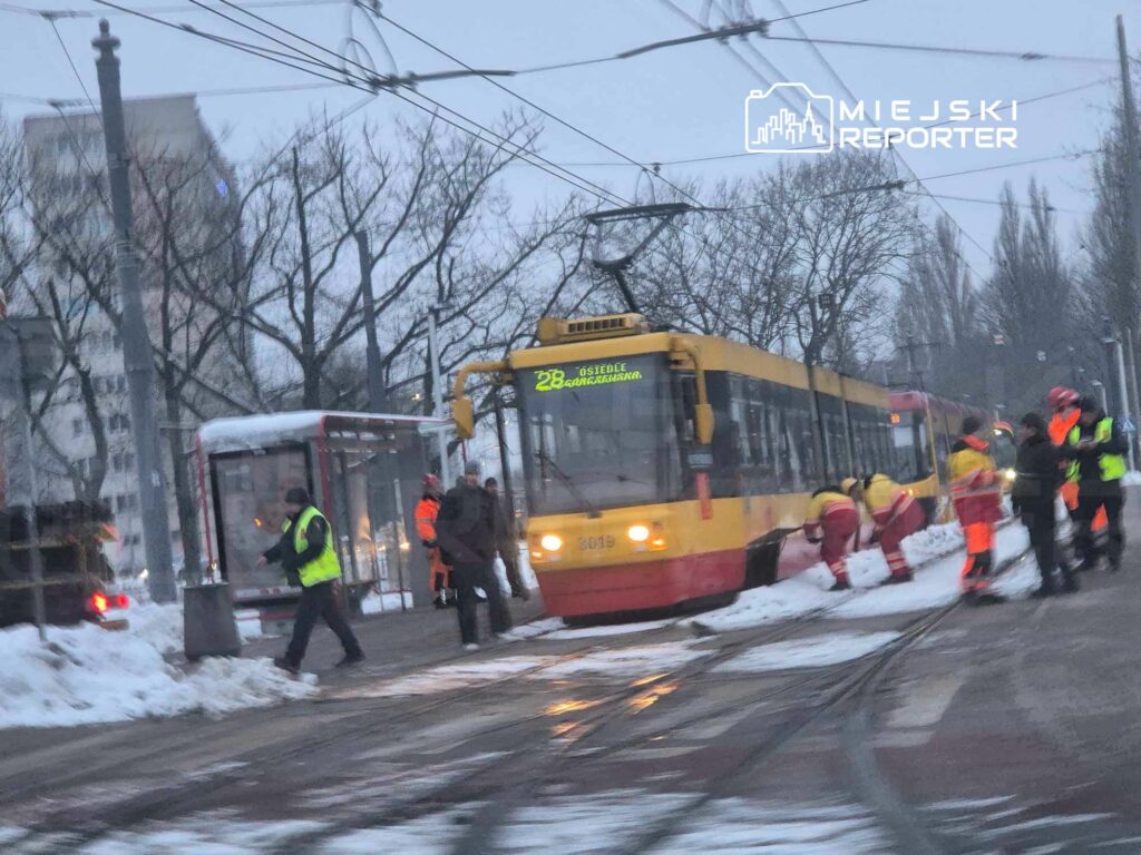 Tramwaj stojący na przystanku, a pracownicy w odblaskowych kamizelkach usuwają śnieg z torowiska.
