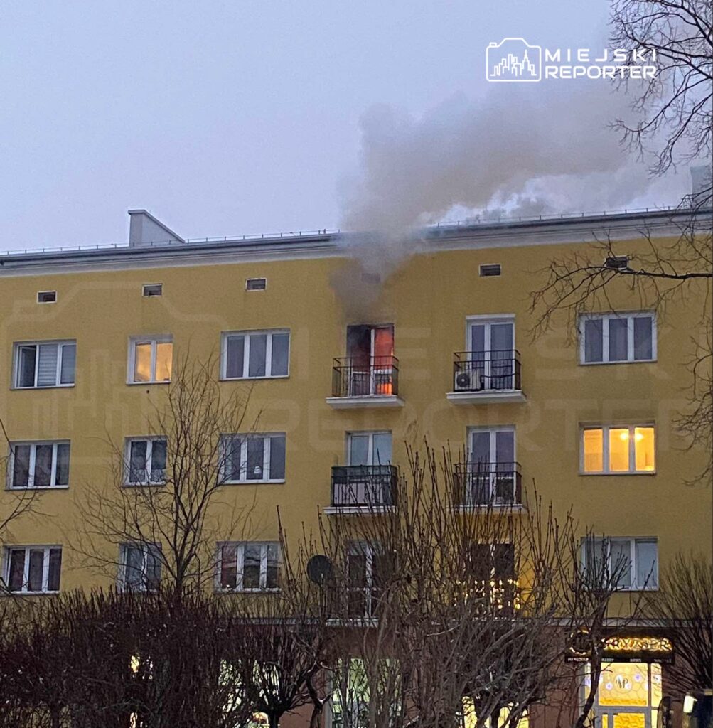 Dym wydobywający się z okna mieszkania na drugim piętrze żółtego budynku mieszkalnego w zimowej scenerii.