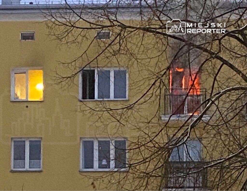 Płomienie wydobywające się z okna mieszkania na żółtej elewacji budynku, otoczone gałęziami drzew.