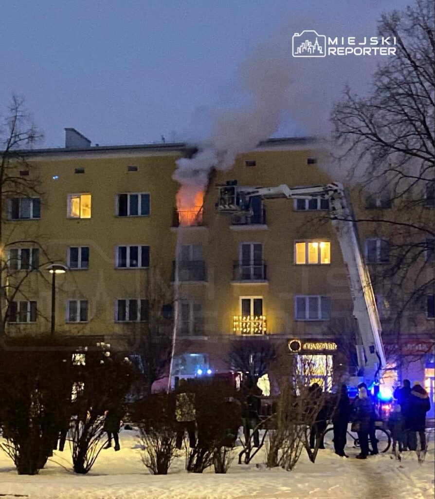 Strażacy gaszą pożar na trzecim piętrze budynku mieszkalnego, otoczeni przez gapiów w zimowej scenerii.