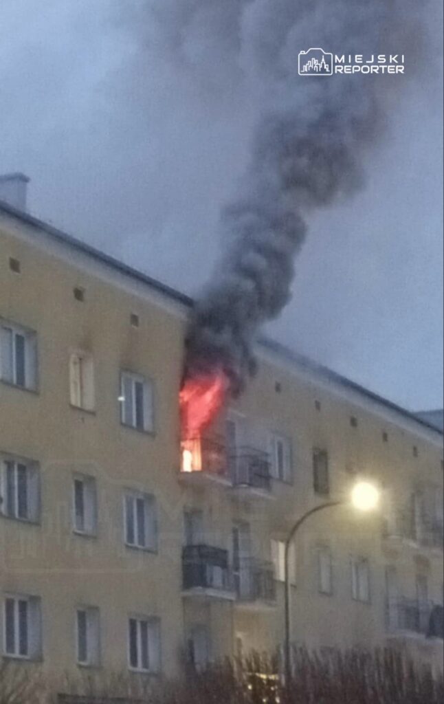 Płomienie wydobywają się z okna mieszkania w bloku, a nad nim unosi się gęsty dym.