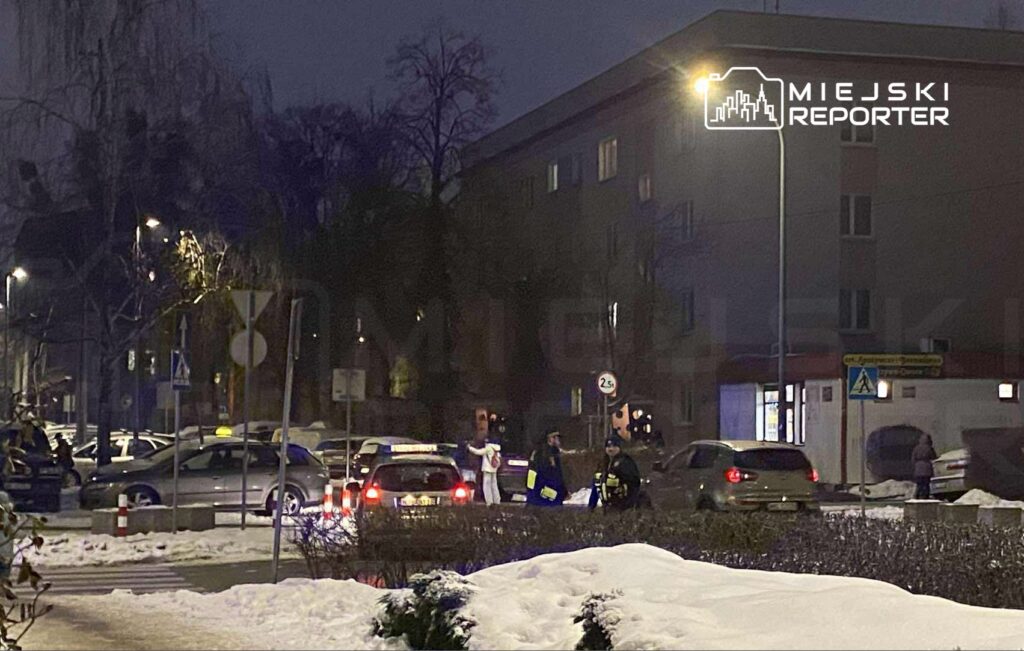 Grupa policjantów i strażaków w nocy interweniuje na oświetlonej ulicy, obok zaparkowanych samochodów.