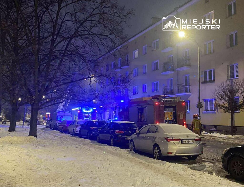 Strażacy pracują przy wozie strażackim na oświetlonej ulicy, pokrytej śniegiem, w nocy.