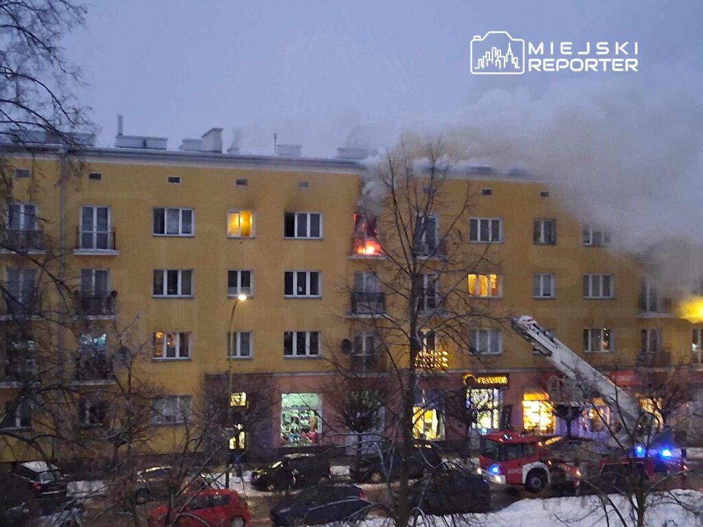 Wielopiętrowy budynek mieszkalny z płonącym oknem, w tle widać dym oraz wóz strażacki z drabiną.