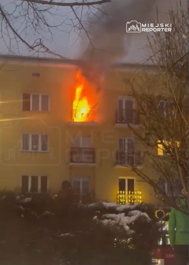 Płomienie wydobywające się z okna na drugim piętrze żółtego budynku mieszkalnego. W tle widać ludzi obserwujących zdarzenie.