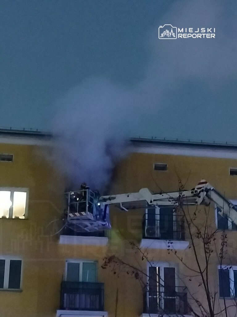 Strażacy na podnośniku gaszą dym wydobywający się z okna budynku mieszkalnego w nocy.