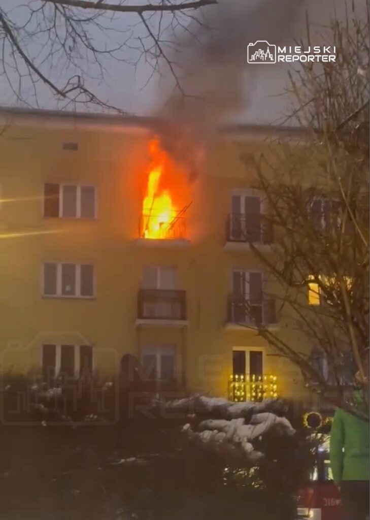 Płomienie wydobywają się z okna na wyższym piętrze żółtego budynku mieszkalnego, otoczonego gałęziami drzew.