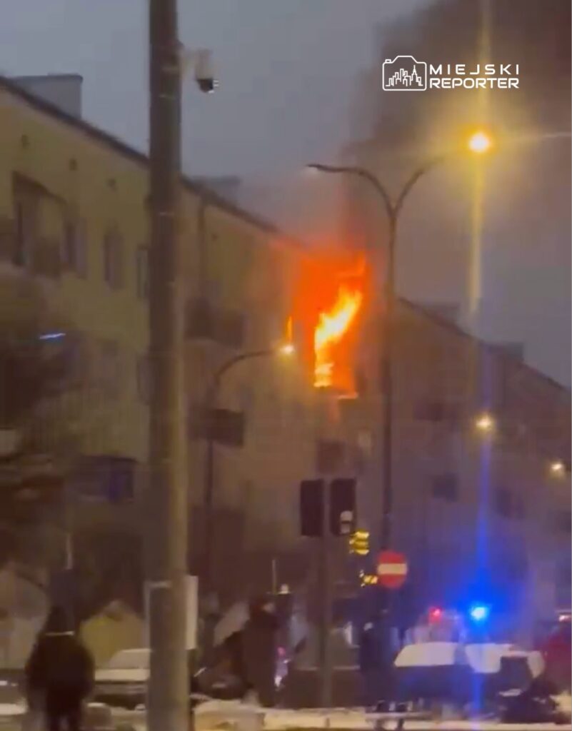 Płomienie wydobywają się z okna budynku mieszkalnego w nocy, wokół zbierają się ludzie oraz pojazdy służb ratunkowych.