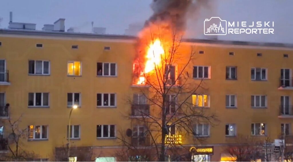 Płomienie wydobywające się z okna mieszkania na trzecim piętrze żółtego budynku wielorodzinnego.