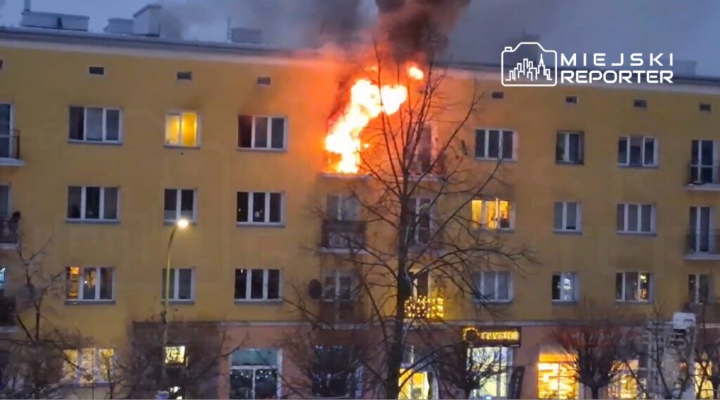 Ogień wydobywający się z okna na trzecim piętrze żółtego budynku mieszkalnego, otoczony drzewami.