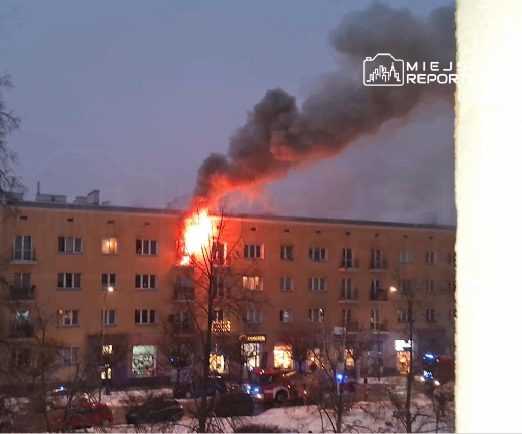 Ogień wydobywający się z okna na czwartym piętrze budynku mieszkalnego, nad którym unosi się gęsty dym. W tle widać pojazdy straży pożarnej.