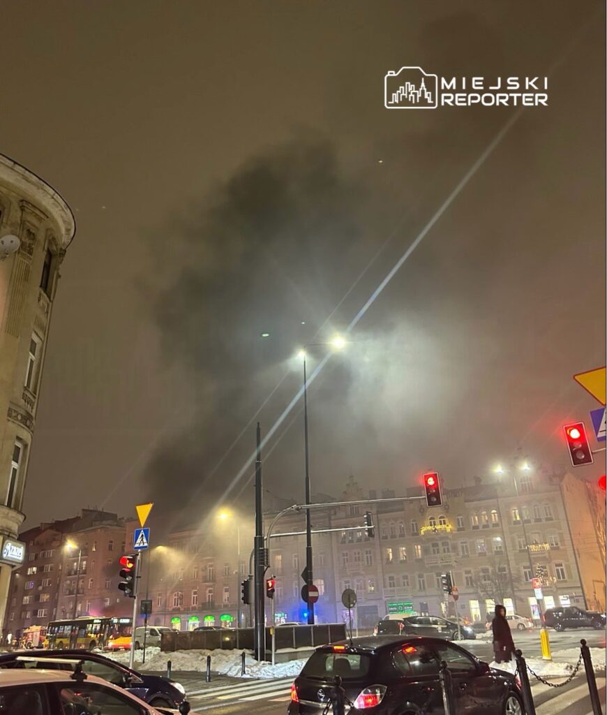 Gęsty dym unosi się nad ulicą w nocy, przy czerwonych sygnalizatorach i oświetlonych budynkach. W tle widoczny autobus.