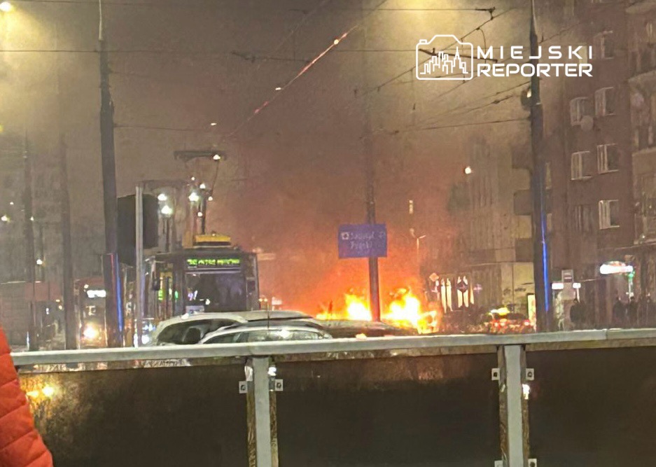 Pojazdy zatrzymane na ulicy w pobliżu ognia i dymu, w tle migające światła miejskie.