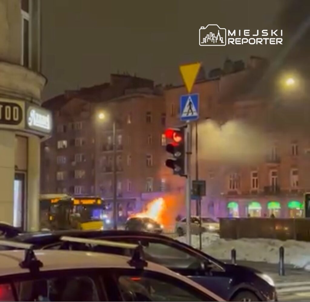 Pojazd płonący na ulicy w pobliżu przystanku autobusowego, w tle widać dym i policyjne światła.