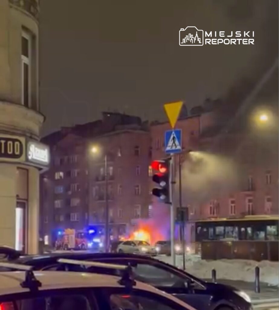 Pożar pojazdu na ulicy, w tle straż pożarna i dym unoszący się z płonącego samochodu.