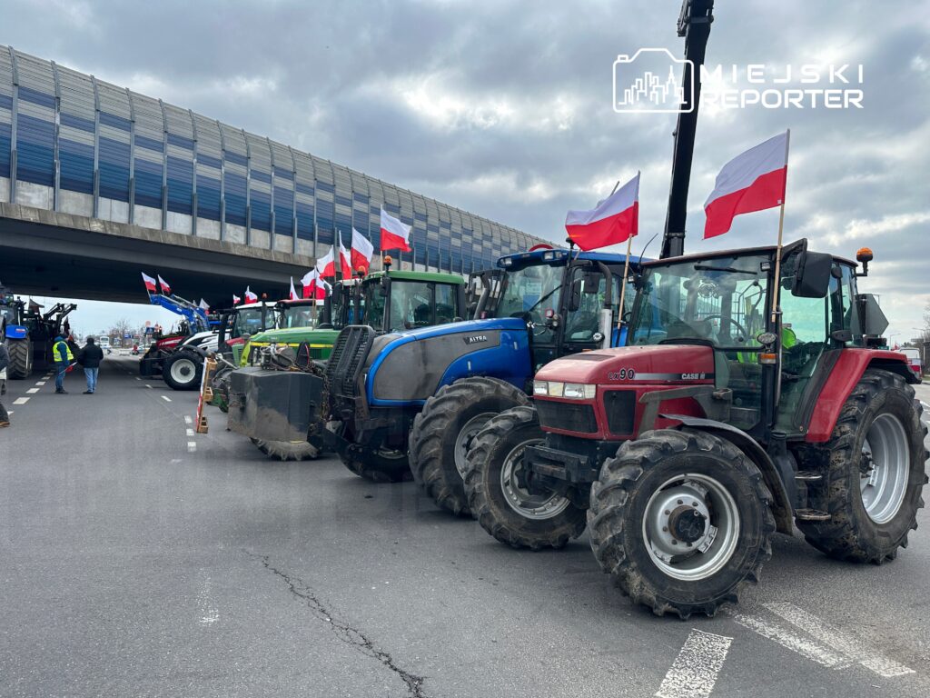 Rolnicy blokują drogę, stojąc obok ciągników z polskimi flagami, pod wiaduktem w pochmurny dzień.