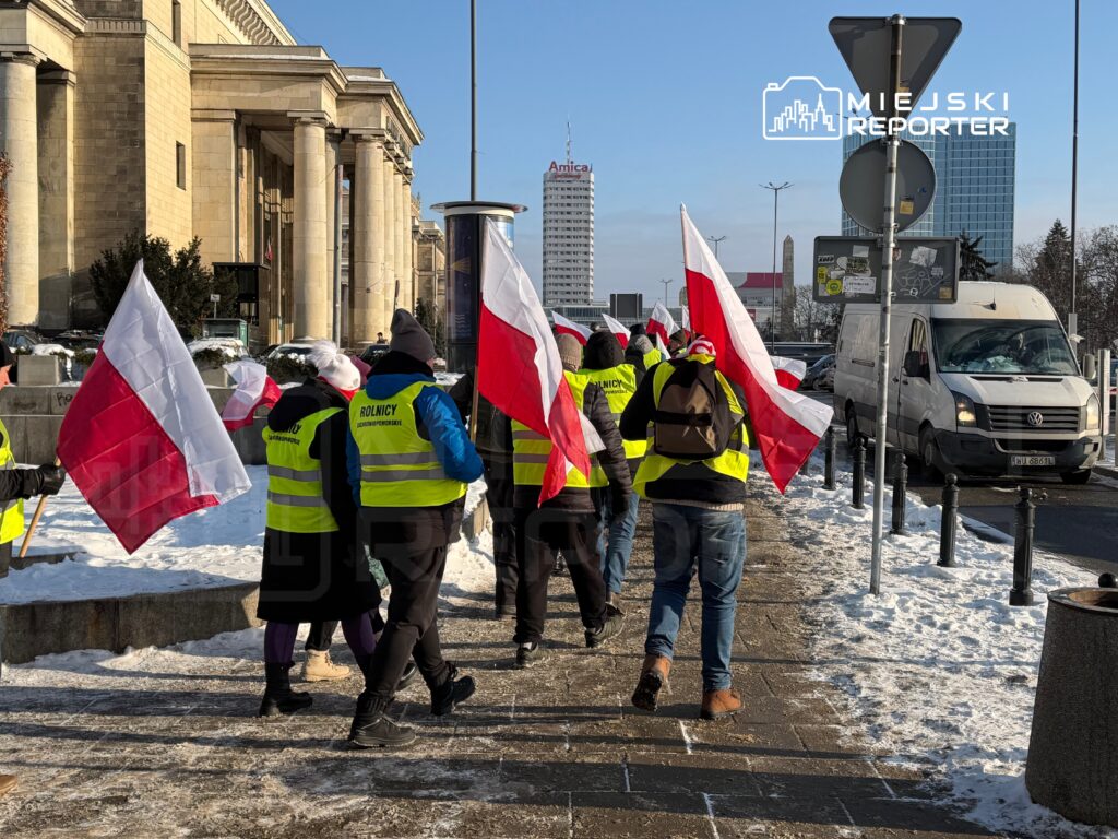 Grupa osób w kamizelkach odblaskowych niesie flagi Polski, przemierzając zaśnieżony chodnik w miejskim otoczeniu.