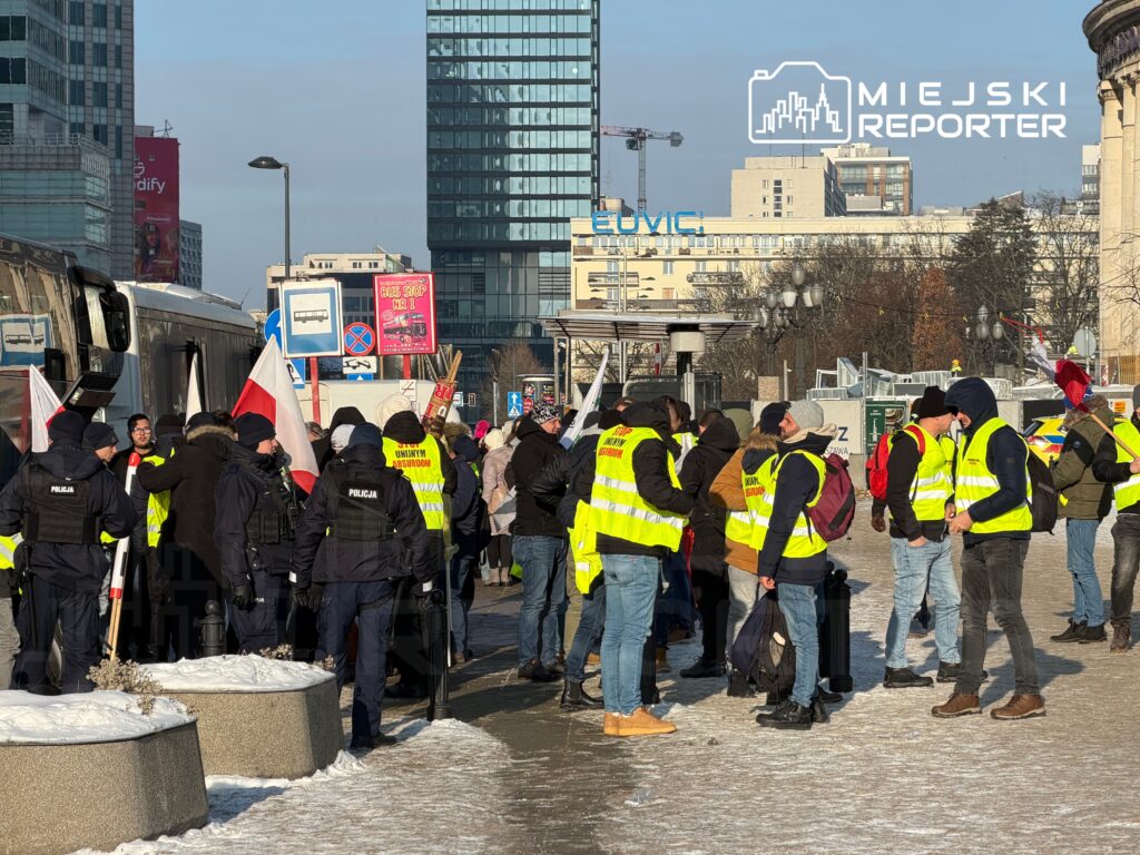 Grupa osób w kamizelkach odblaskowych i mundurach policji gromadzi się na ulicy, trzymając flagi.