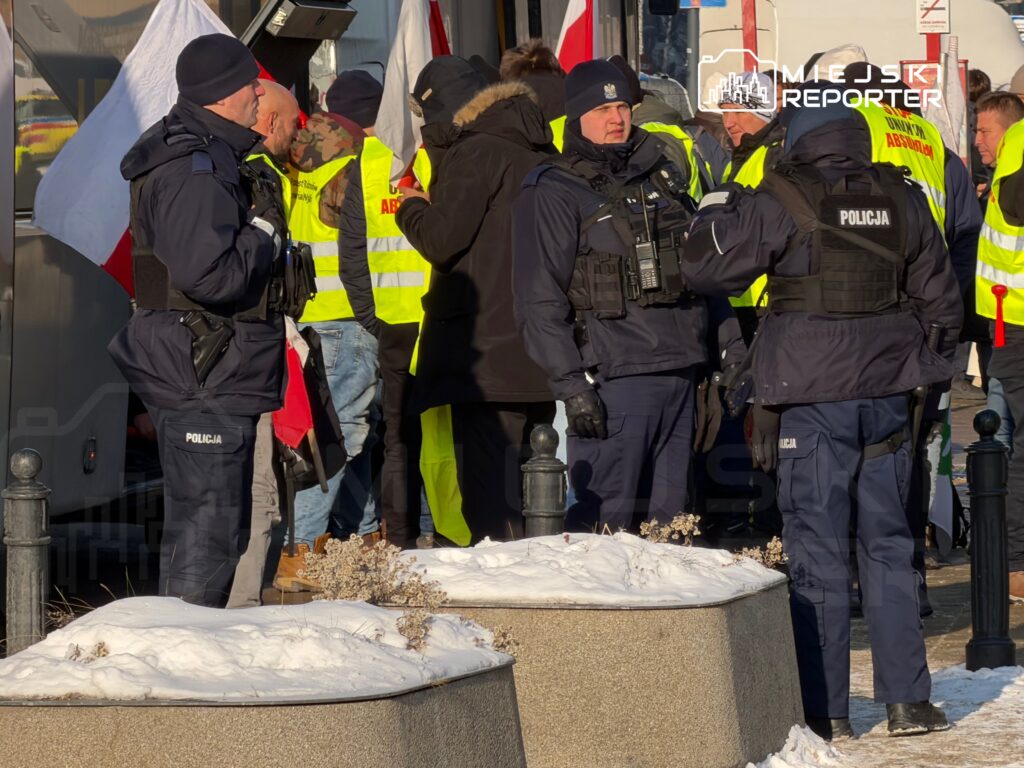 Grupa policjantów w mundurach rozmawia z uczestnikami protestu, którzy noszą odblaskowe kamizelki. W tle widoczne flagi.