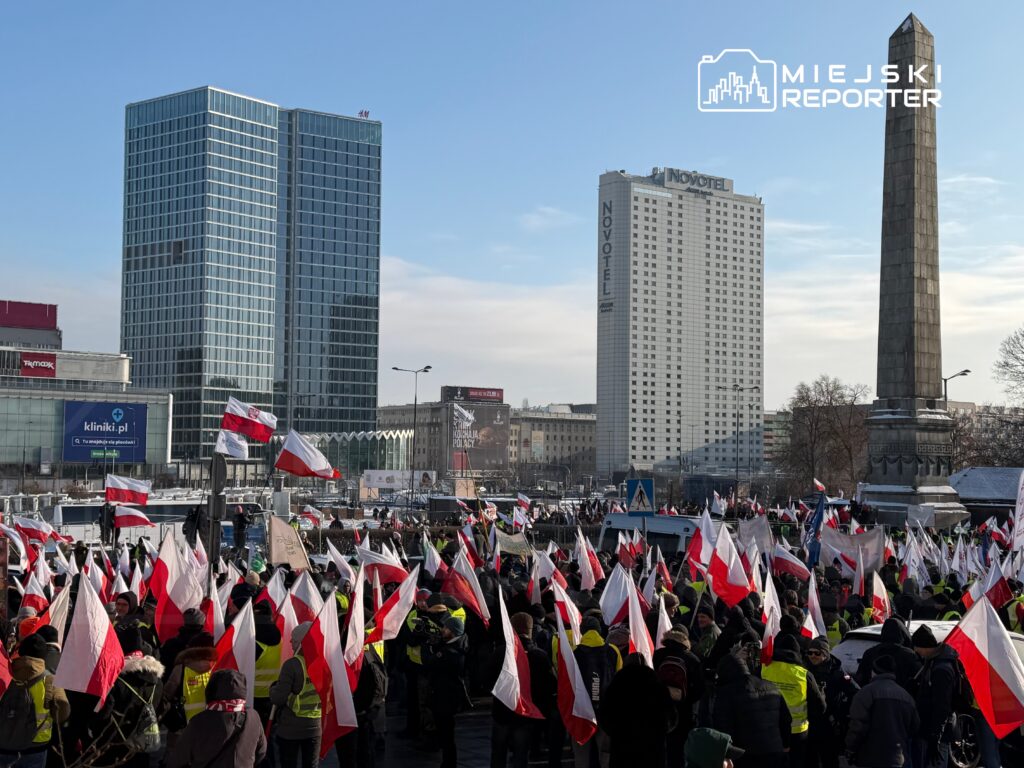 Tłum ludzi w żółtych kamizelkach i z flagami narodowymi protestuje na placu w centrum miasta, w tle nowoczesne budynki.